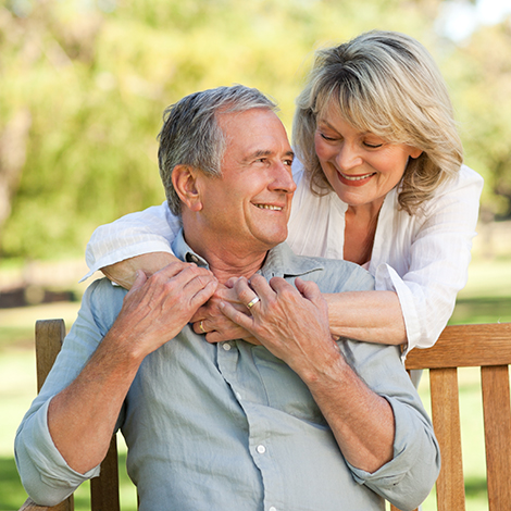 middle aged couple, man sitting on bench and woman standing behind with arms around man's shoulders smiling