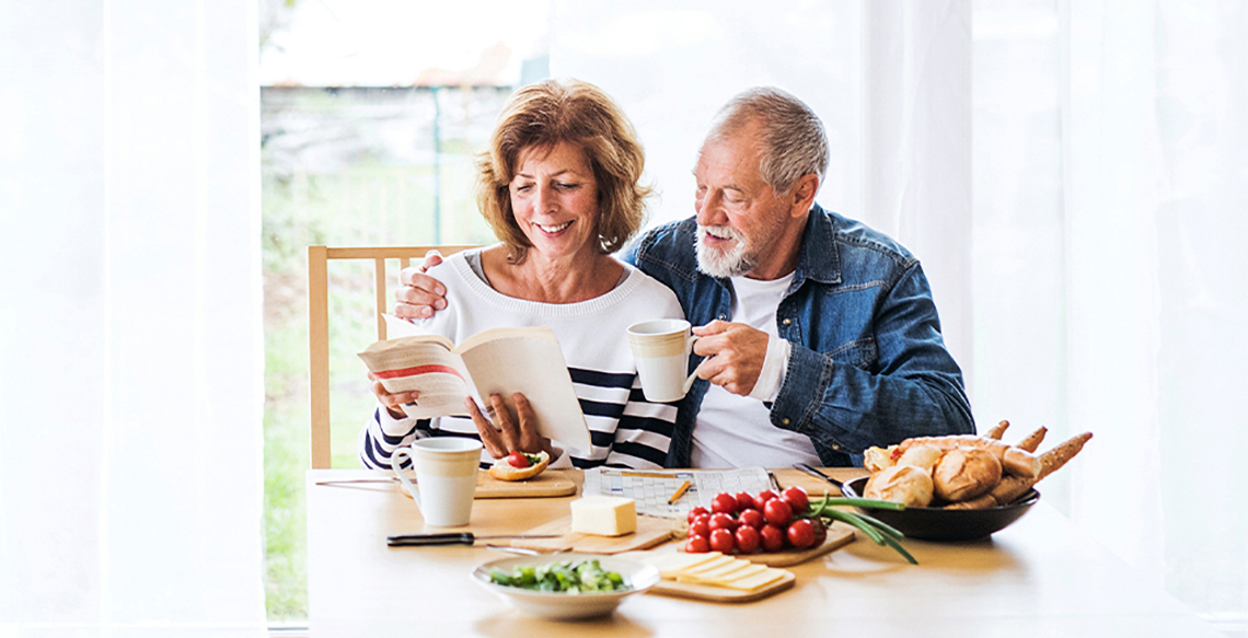 Man and woman sitting at kitchen table together. Woman is reading a book and the man is holding a coffee cup. Their is food on the table.