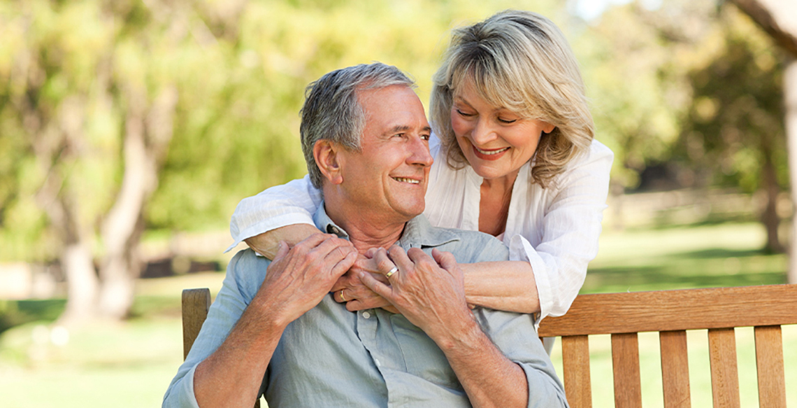 Man sitting on a bench outside and woman hugging him from behind.